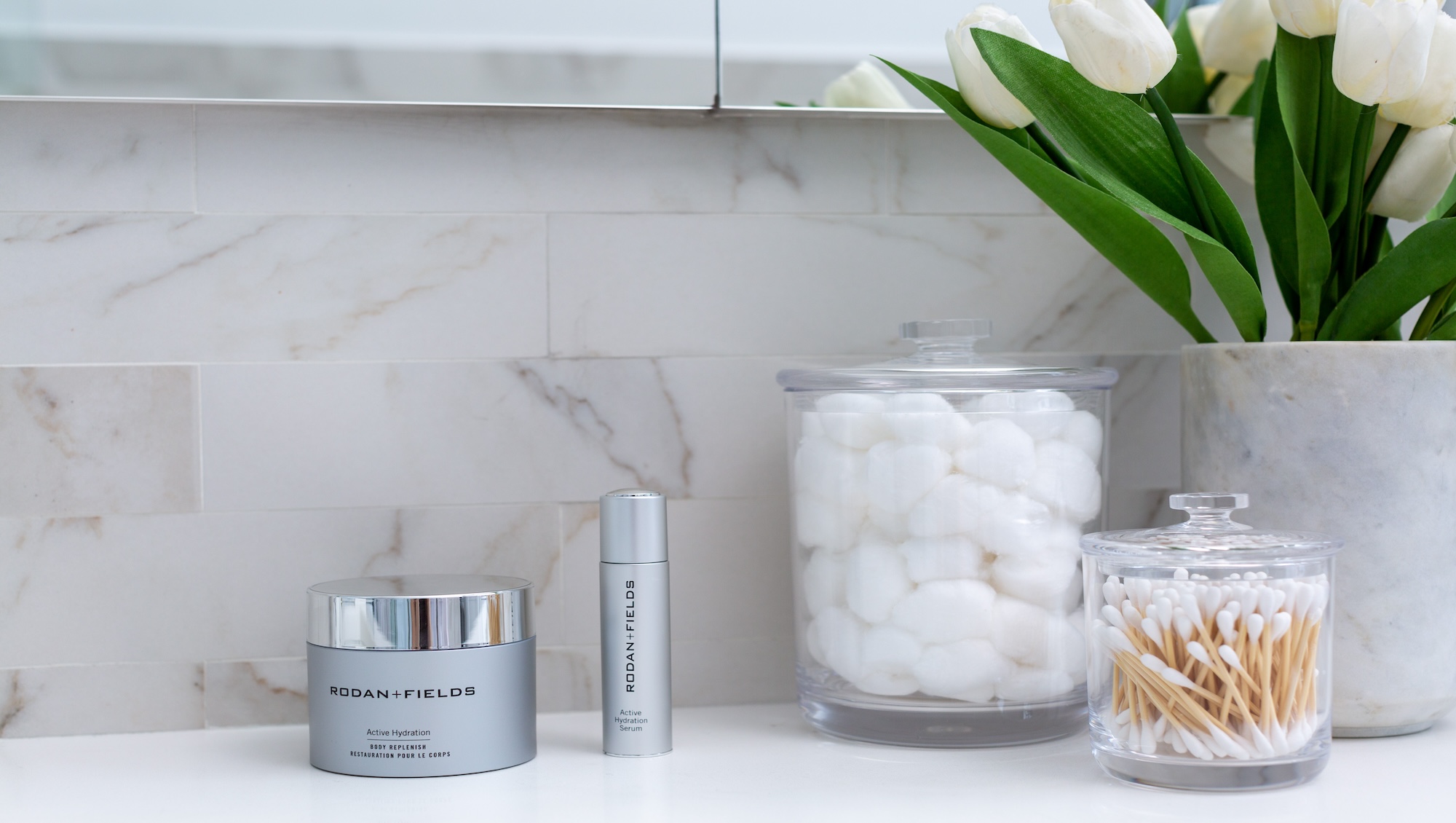 Skincare from Rodan + Fields next to cotton balls and Q-tips arranged on a bathroom counter.
