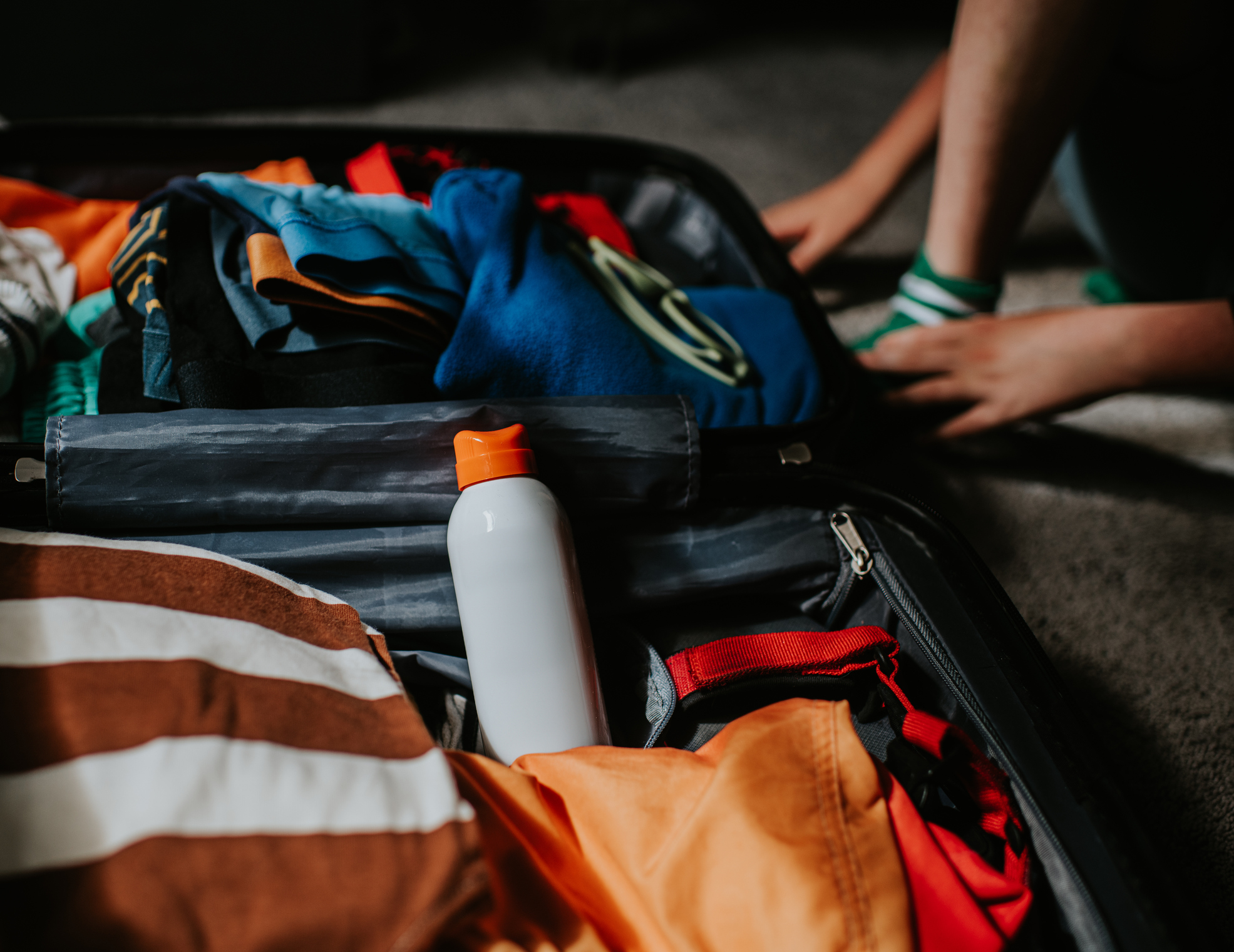 A spray bottle of unbranded sun cream amongst the colourful contents of a child's packed suitcase, ready for a vacation in the sun