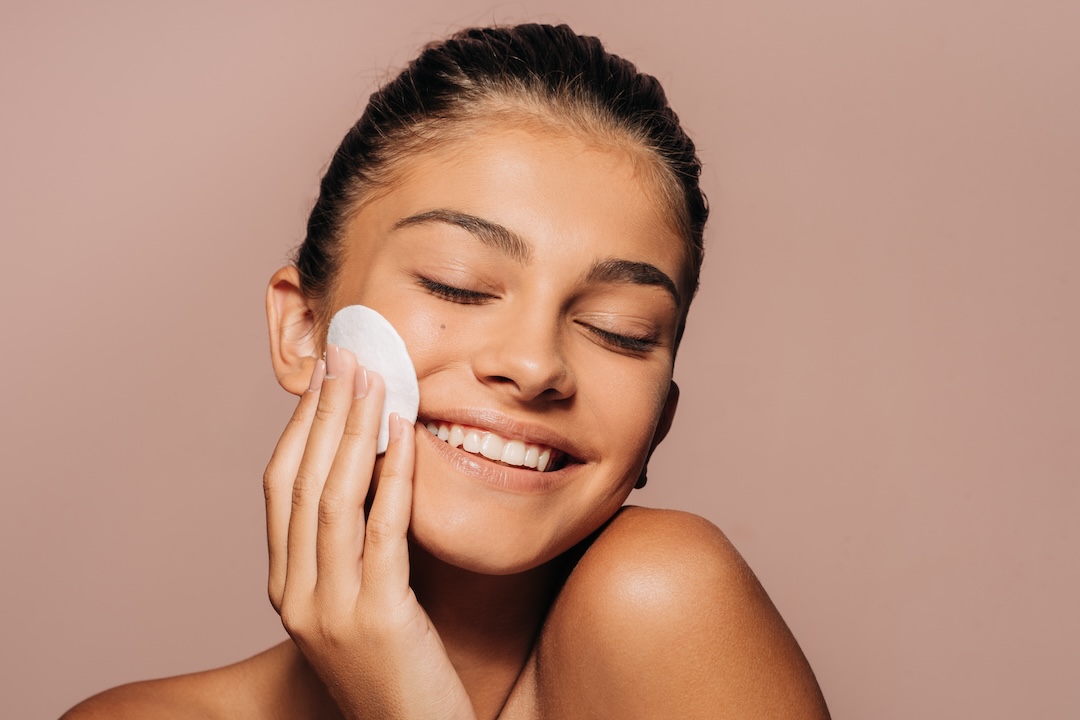 This is a close-up photo of a smiling young woman with glowing skin. She is holding soft cotton pads, one covering her eye and the other touching her cheek. Her hair is tied back, and the background is a soft pink color. The image is studio taken and it is showing a skincare routine.