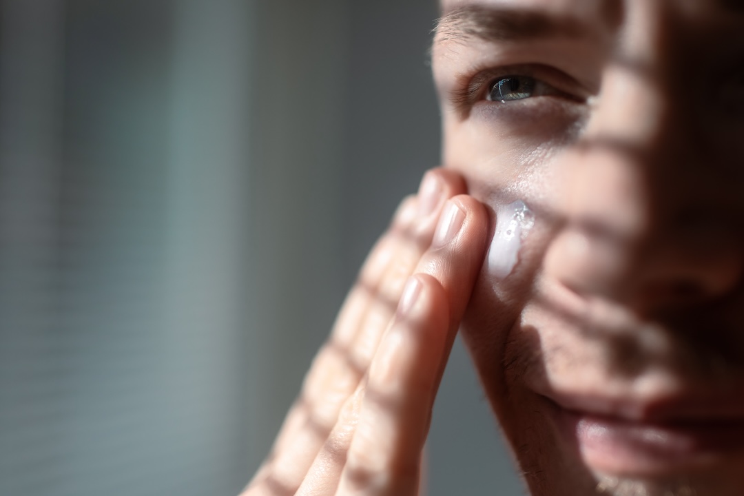 Men skin care routine. Close-up applying cream on face. Sunny light with shadows of blinds.