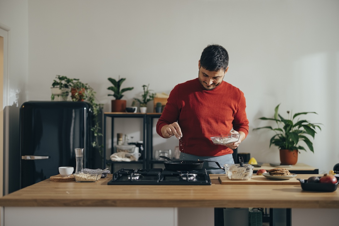 Handsome cheerful man preparing a meal in the kitchen. He is standing wearing a red sweater and adding salt to a pan with his fingers.