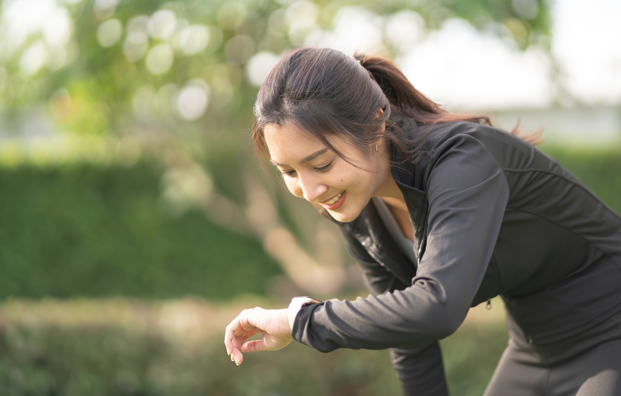 Young Asian sports woman resting after working out outdoors. Checking pulse and training performance on fitness app on her smartwatch. Health and fitness training with technology.