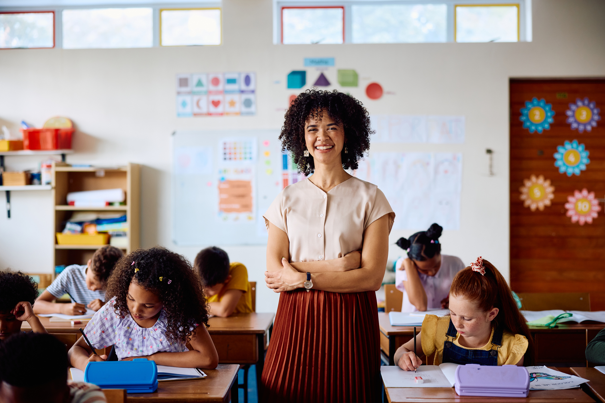 Female teacher in her 20s standing in classroom with arms crossed, looking at camera with cheerful expression