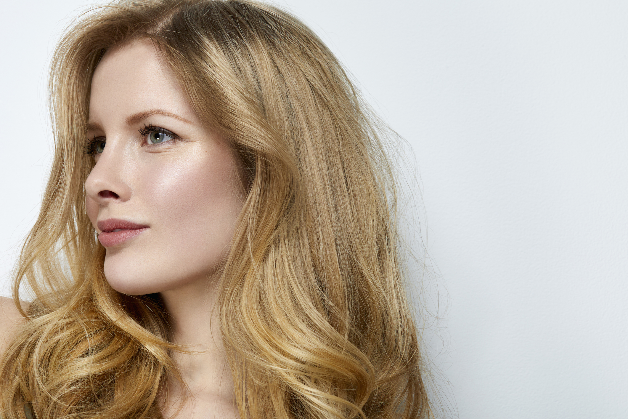 Studio shot of young beautiful woman in front of a white wall. Professional make-up and hairstyle with long blond wavy hair