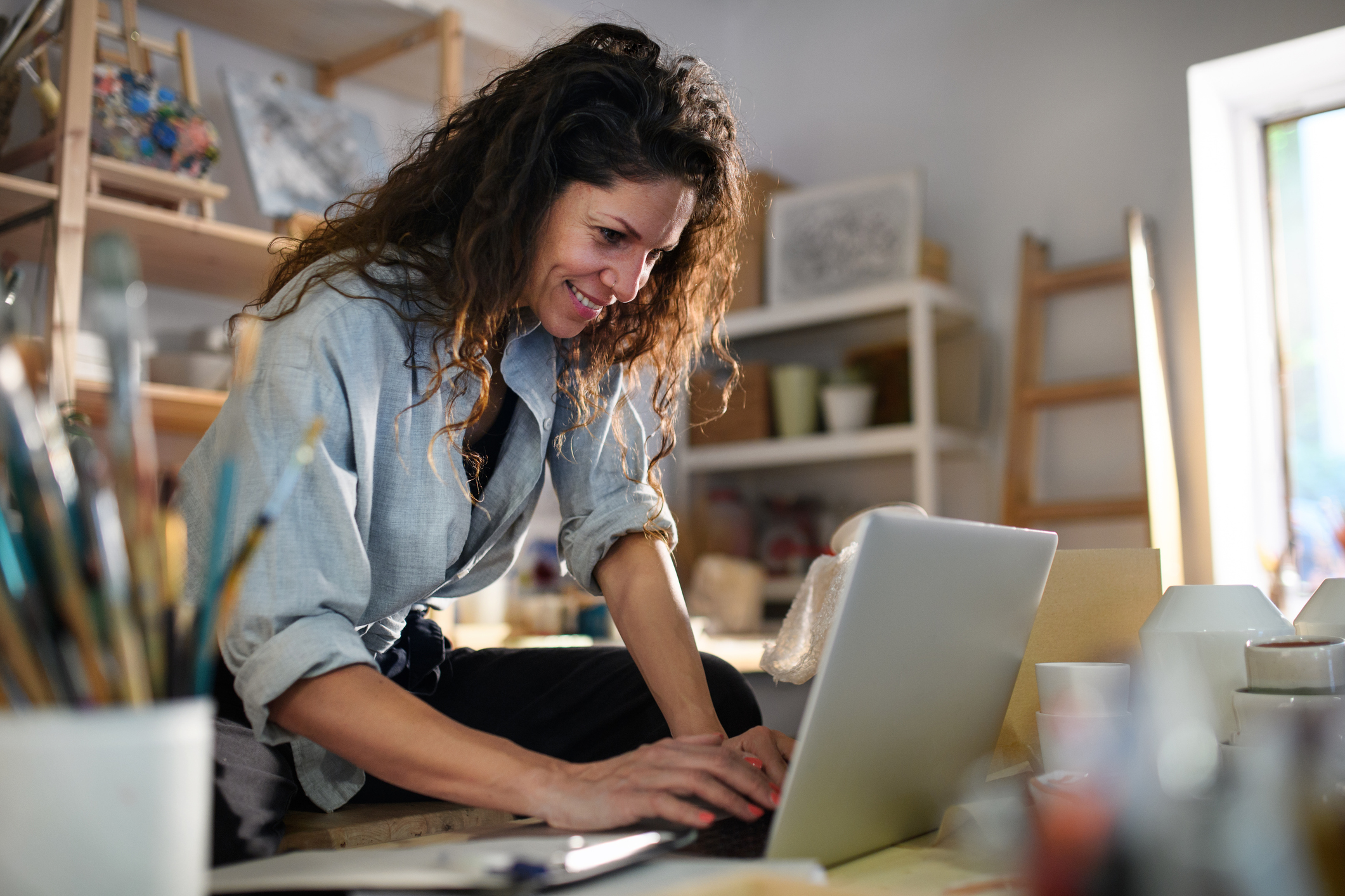 Happy young woman with laptop handles orders in pottery workshop.