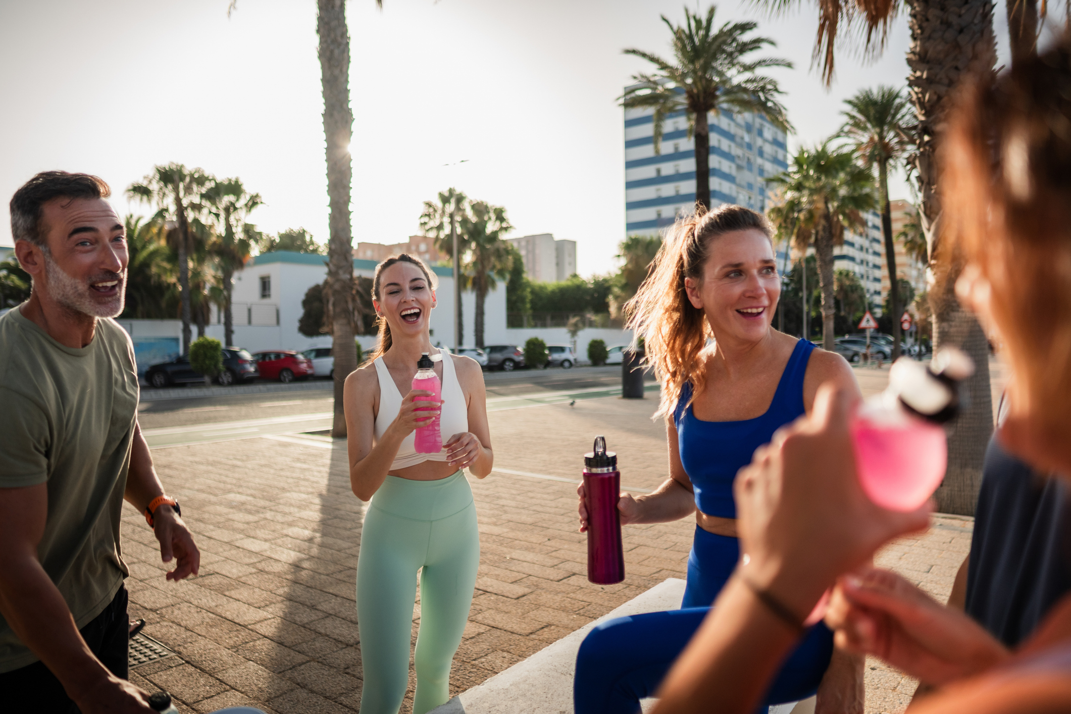 Group of cheerful friends in sportswear laughing and drinking water after exercising outdoors in summer