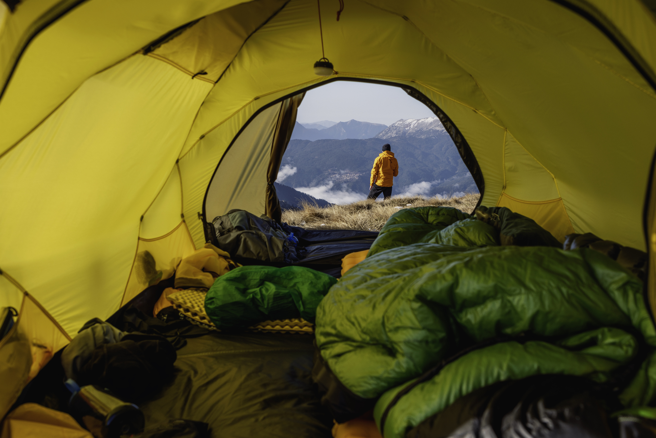A traveler stands outside a tent gazing at mountains during sunrise.