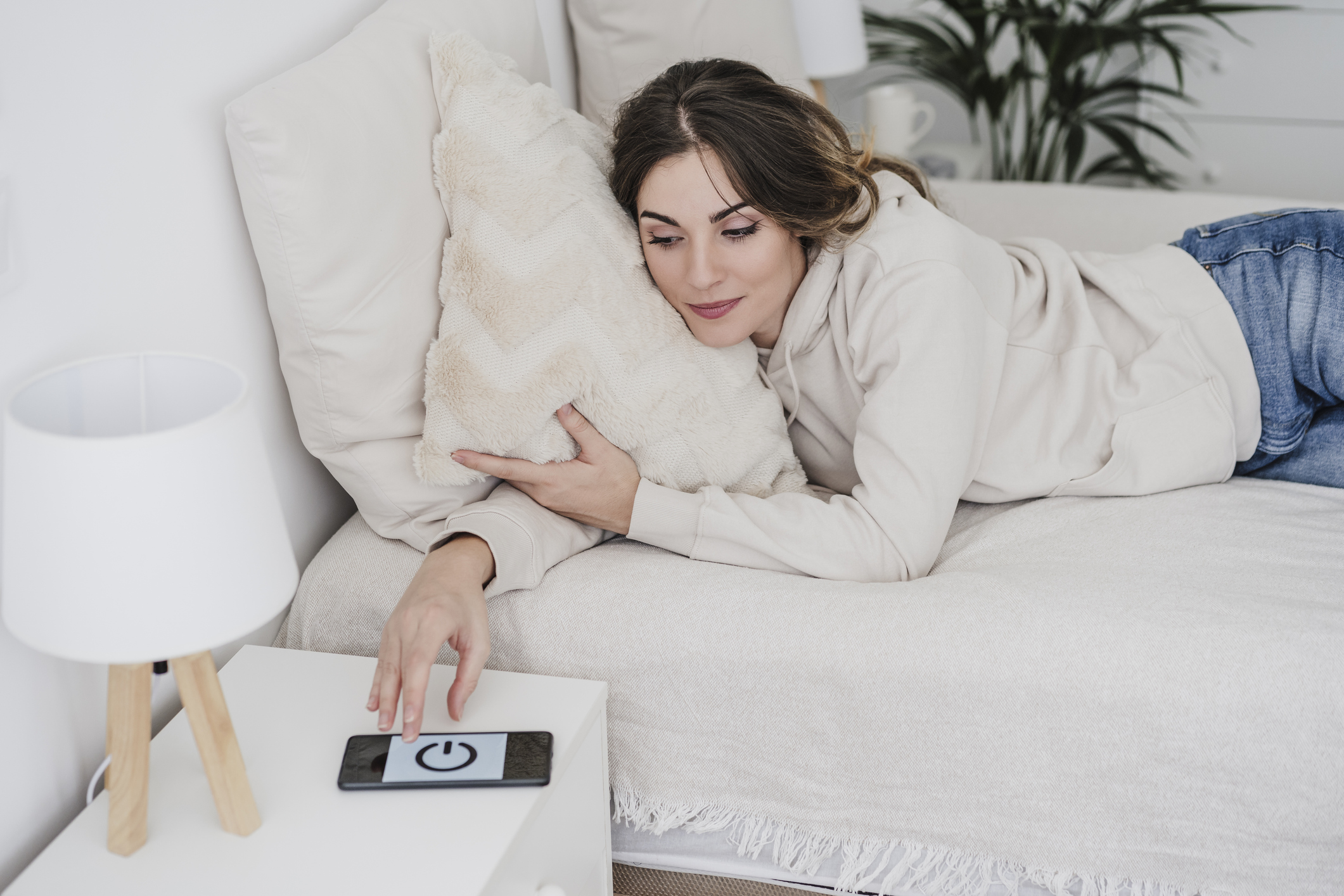 Young woman lying on bed switching off smart phone alarm at home