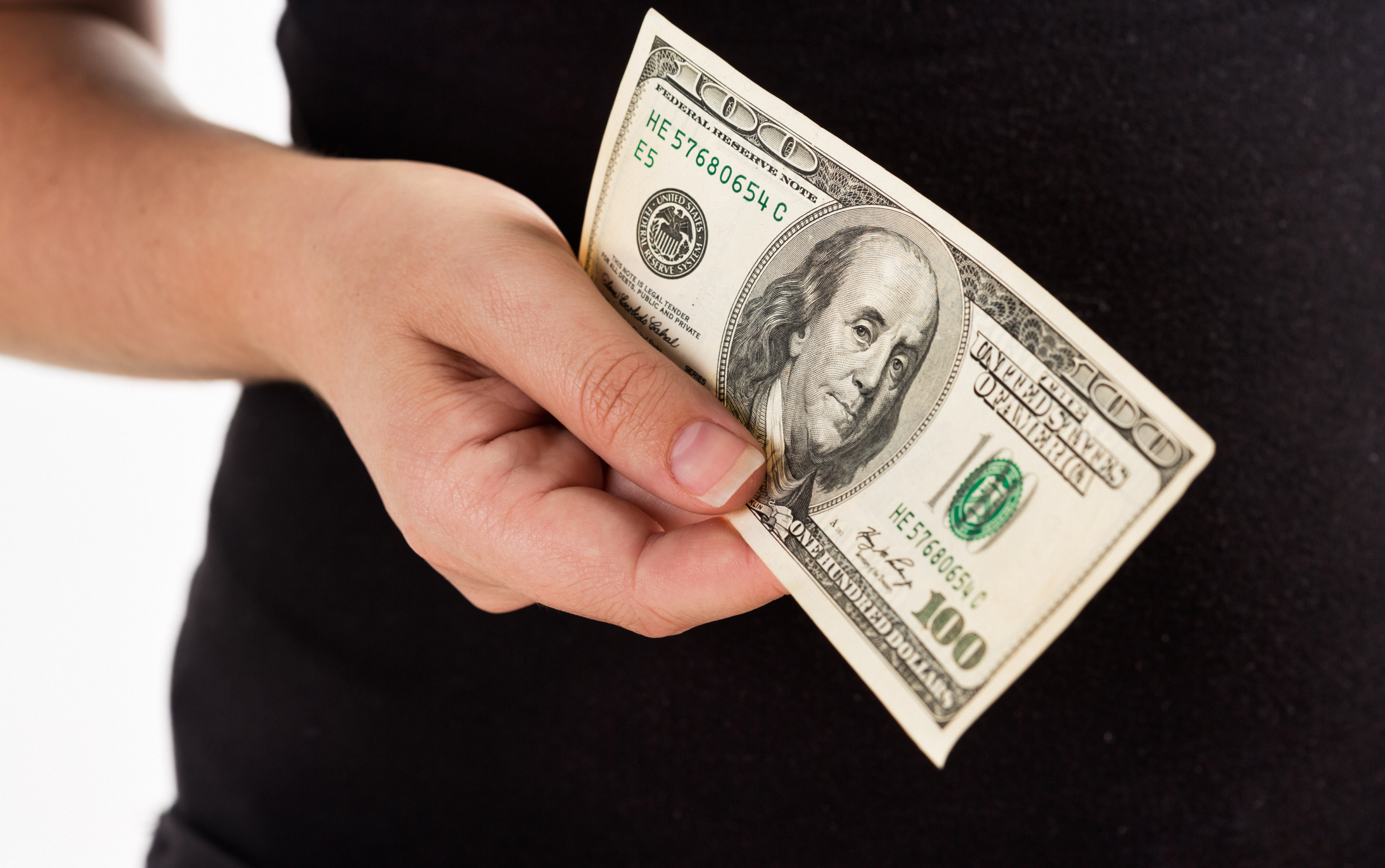 A hand holding a hundred-dollar banknote diagonally against black clothing, with Benjamin Franklin’s portrait clearly visible.