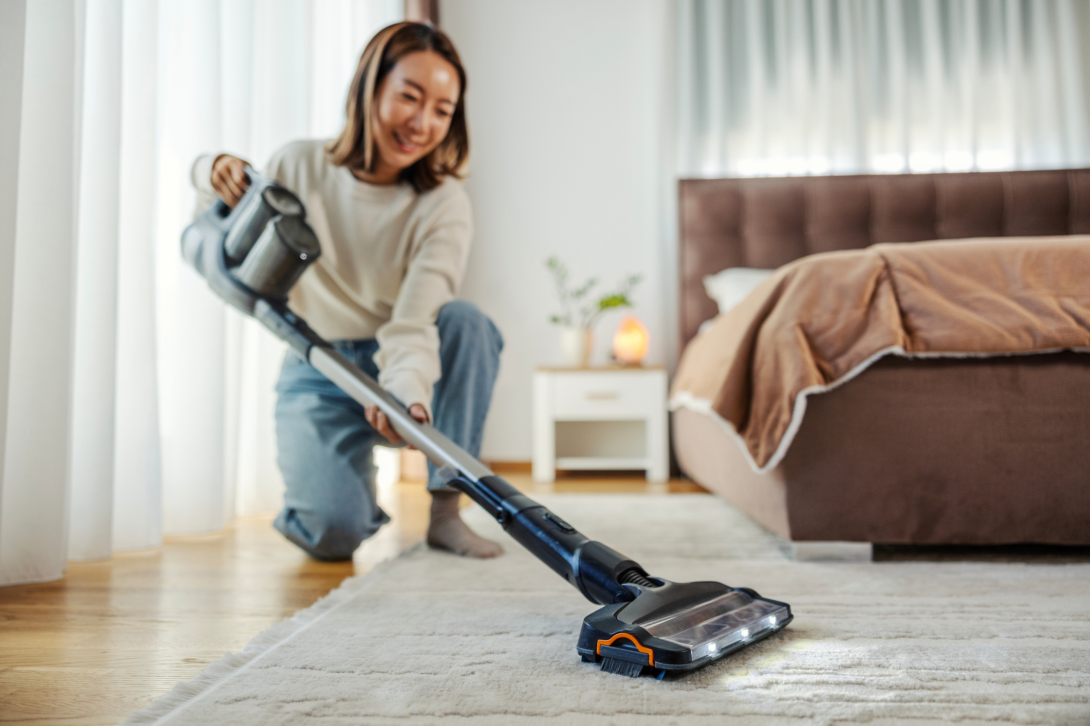Young asian woman vacuuming carpet in her bedroom, smiling, performing daily household chores with a modern appliance