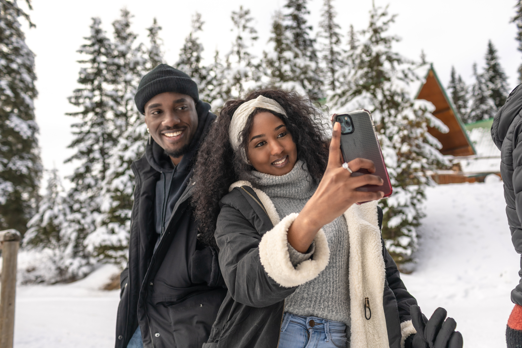 An attractive and stylish couple, dressed in warm outdoor winter apparel pose for a selfie with the woman's smartphone. It's a winter setting.