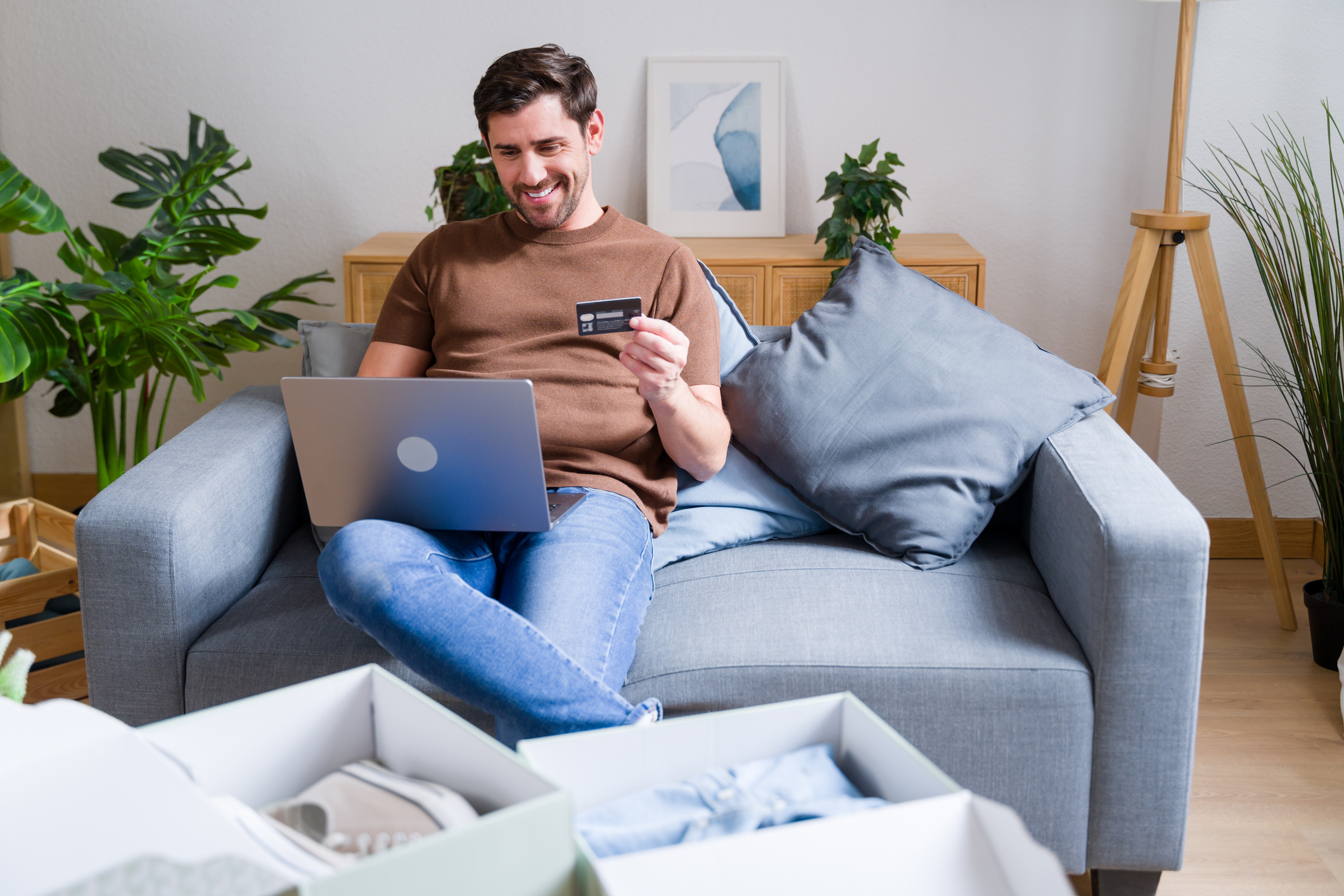 Smiling man is making an online purchase using his laptop and credit card, surrounded by shopping boxes at home