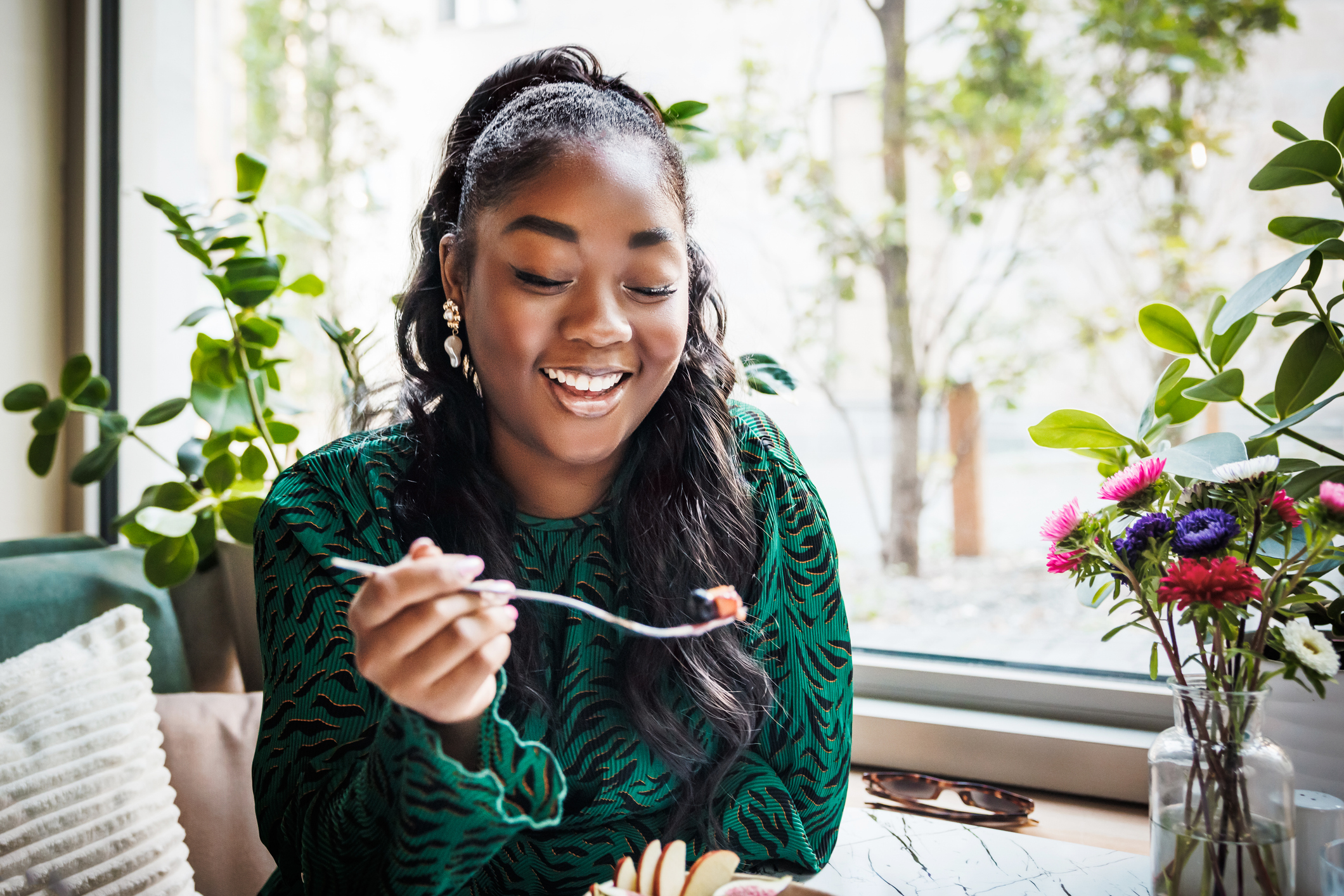 A smiling young woman is captured in a candid moment as she enjoys her food, surrounded by a cozy café atmosphere with lush greenery and vibrant flowers.
