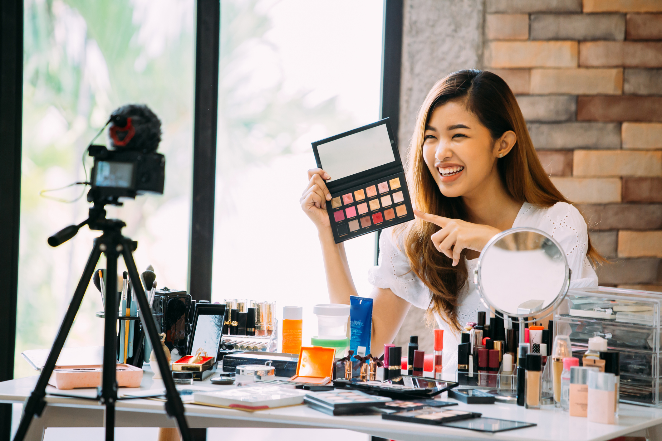 A woman sitting at table and making a video about cosmetics while filming with camera on tripod