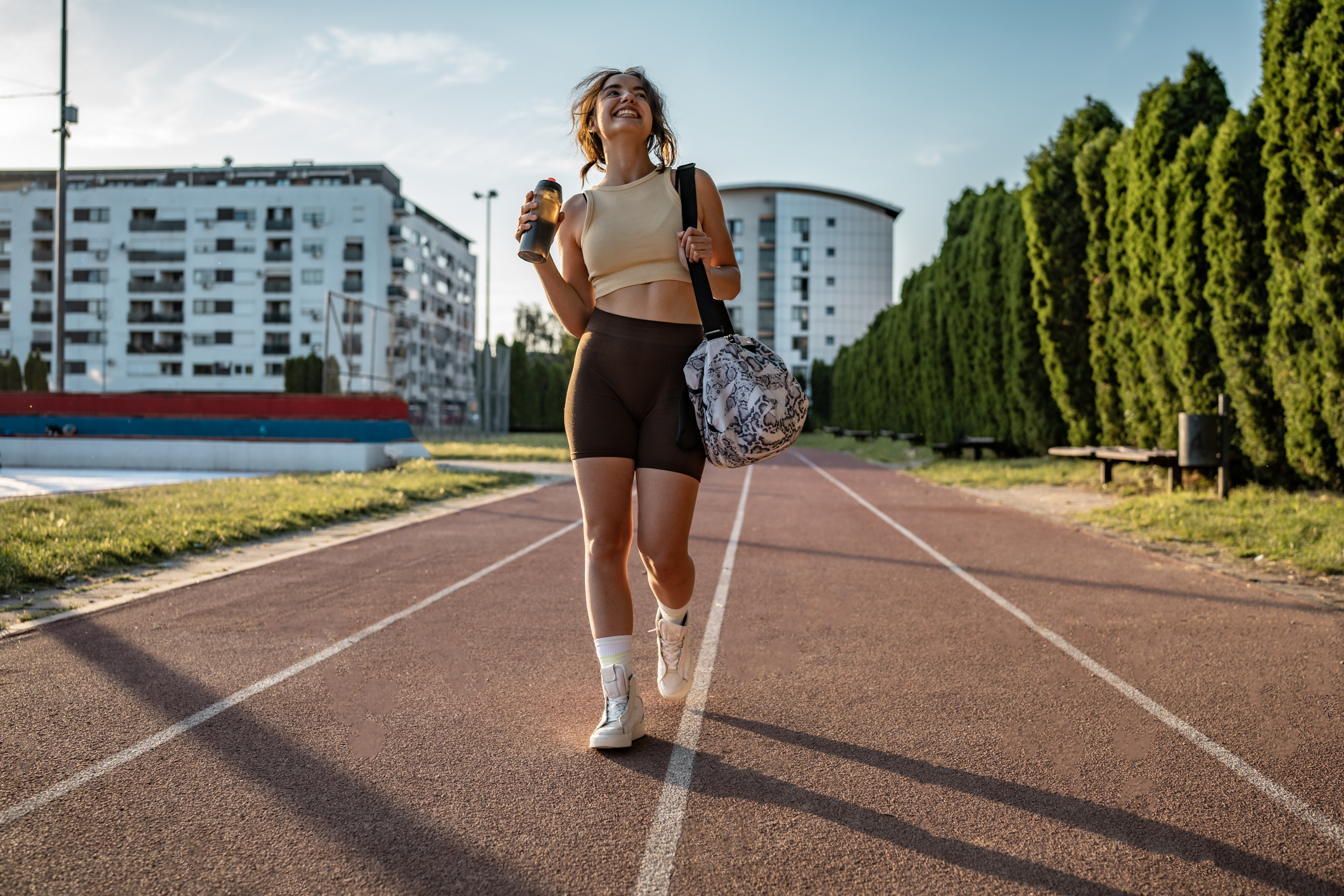 A cheerful woman walking on a running track outdoors, holding a bottle and a bag, enjoying the sunny day.
