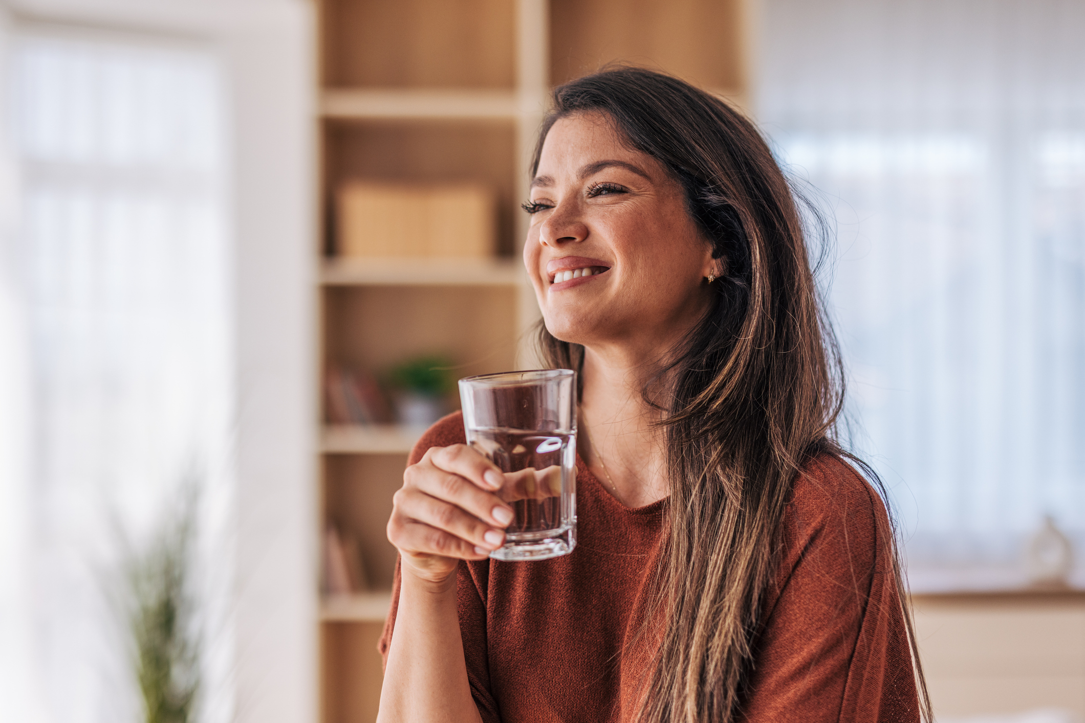 Smiling woman holding a glass of water, enjoying a refreshing drink in the comfort of her home, promoting a healthy lifestyle and hydration