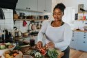 Smiling young woman preparing salad bowl in kitchen at home