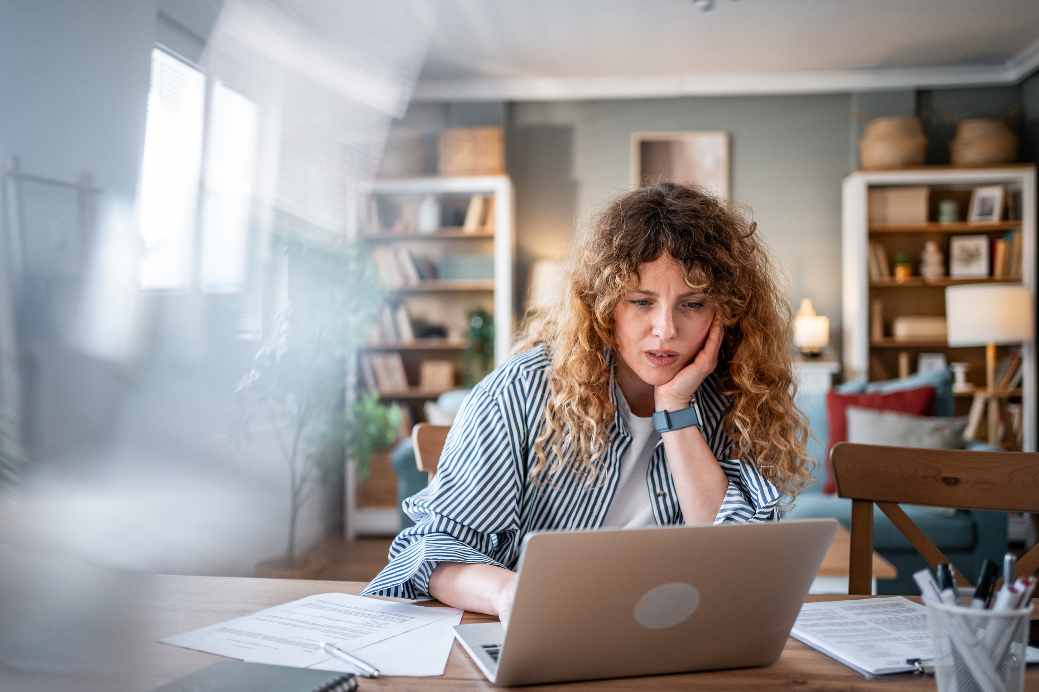 Young adult woman with curly hair looking concerned and thoughtful while using a laptop at a wooden table in her apartment, working remotely and managing her freelance projects
