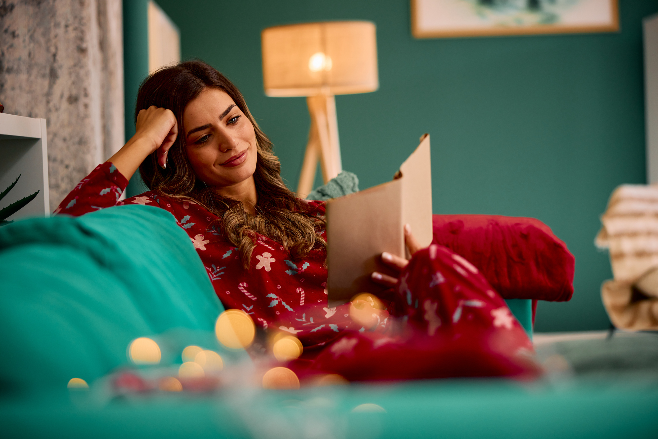 Young woman wearing Christmas pajamas relaxing on the sofa reading a book in a cozy living room with Christmas lights during winter holidays