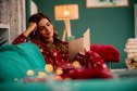 Young woman wearing Christmas pajamas relaxing on the sofa reading a book in a cozy living room with Christmas lights during winter holidays
