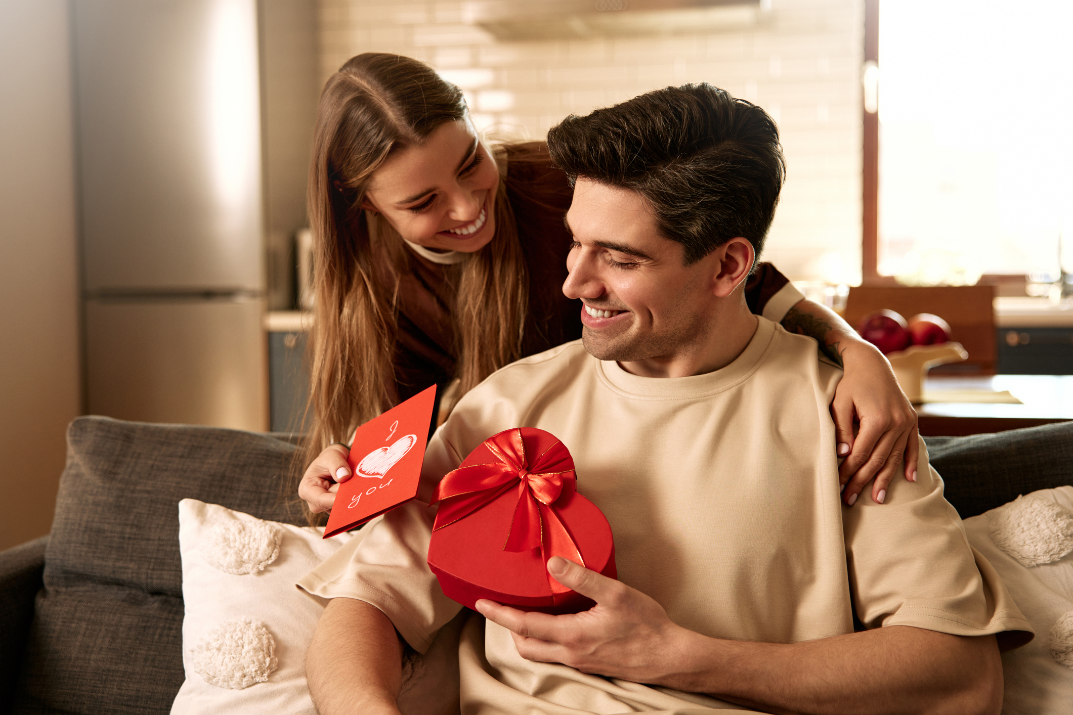 Smiling couple enjoying Valentine's Day at home. Woman giving a red greeting card and hugging man holding heart-shaped gift box. Concept of love, surprise and romantic relationship.