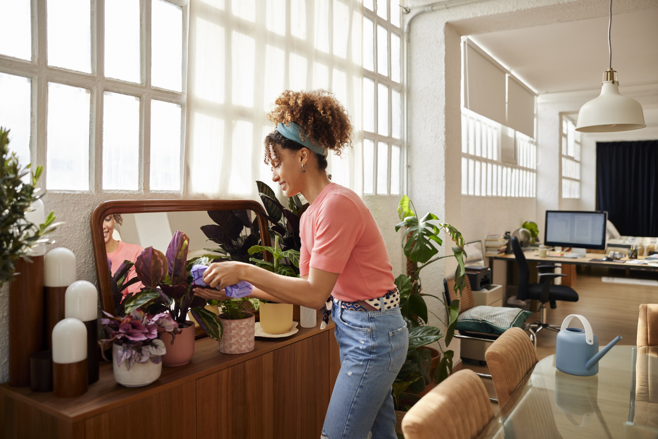 Side view of young woman cleaning leaves of plotted plants at home. Woman with curly hair gardening at home. She is wearing casuals.