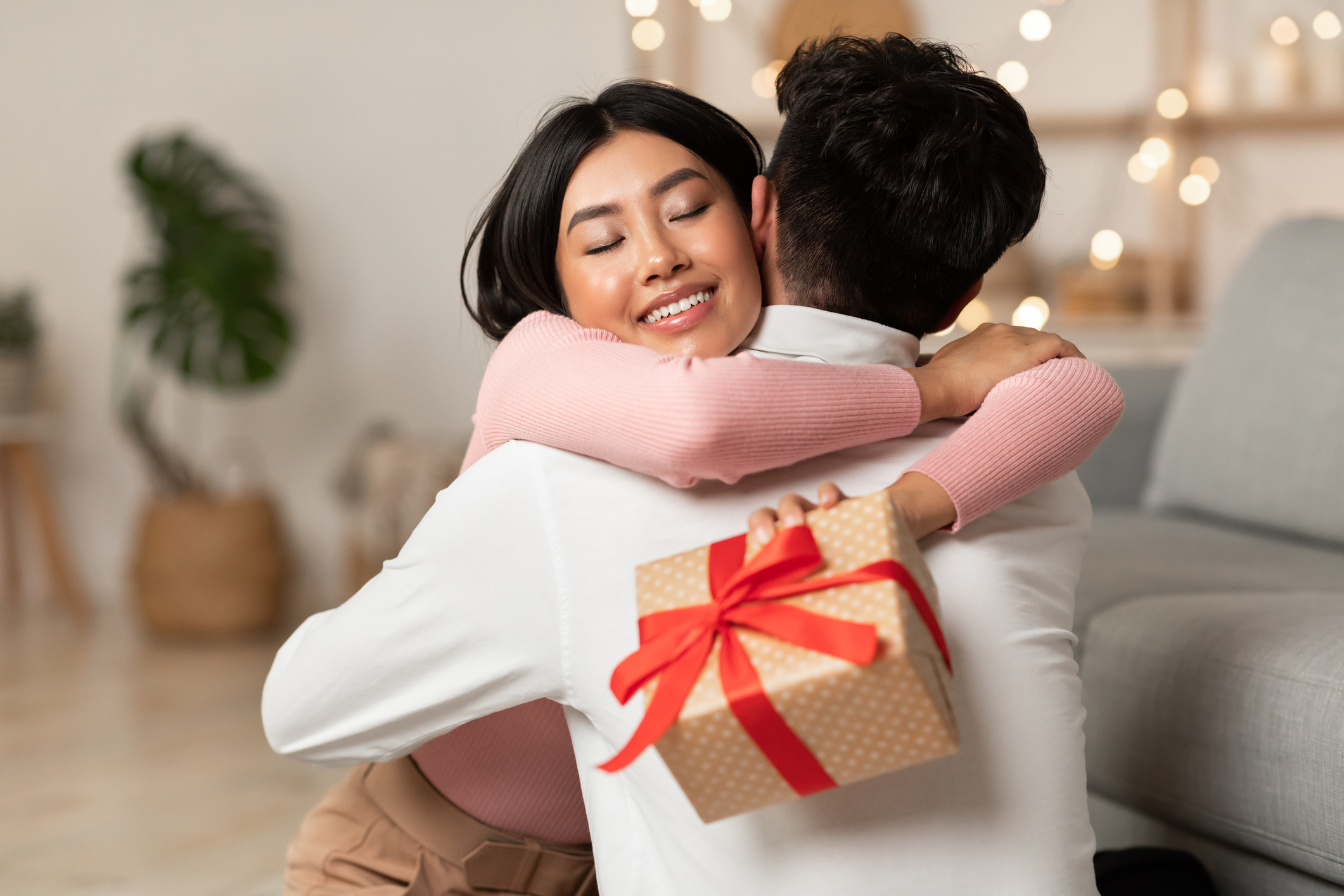 Joyful Wife Embracing Husband Holding Christmas Gift Receiving Presents On Xmas Holiday Or Birthday Sitting At Home. Valentine's Day And Winter Holidays Celebration Concept