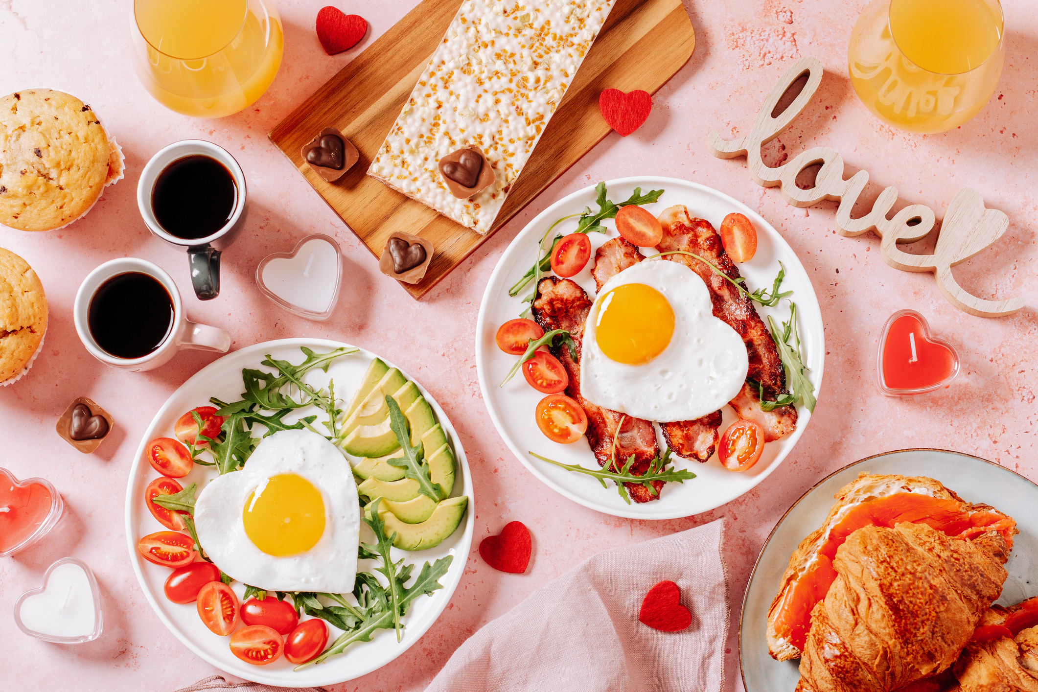 Fried eggs in heart shape, bacon, avocado and vegetables on two white plates, coffee cups and muffins- couple breakfast on Valentines Day or honeymoon on pink background, toned image