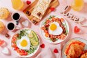 Fried eggs in heart shape, bacon, avocado and vegetables on two white plates, coffee cups and muffins- couple breakfast on Valentines Day or honeymoon on pink background, toned image