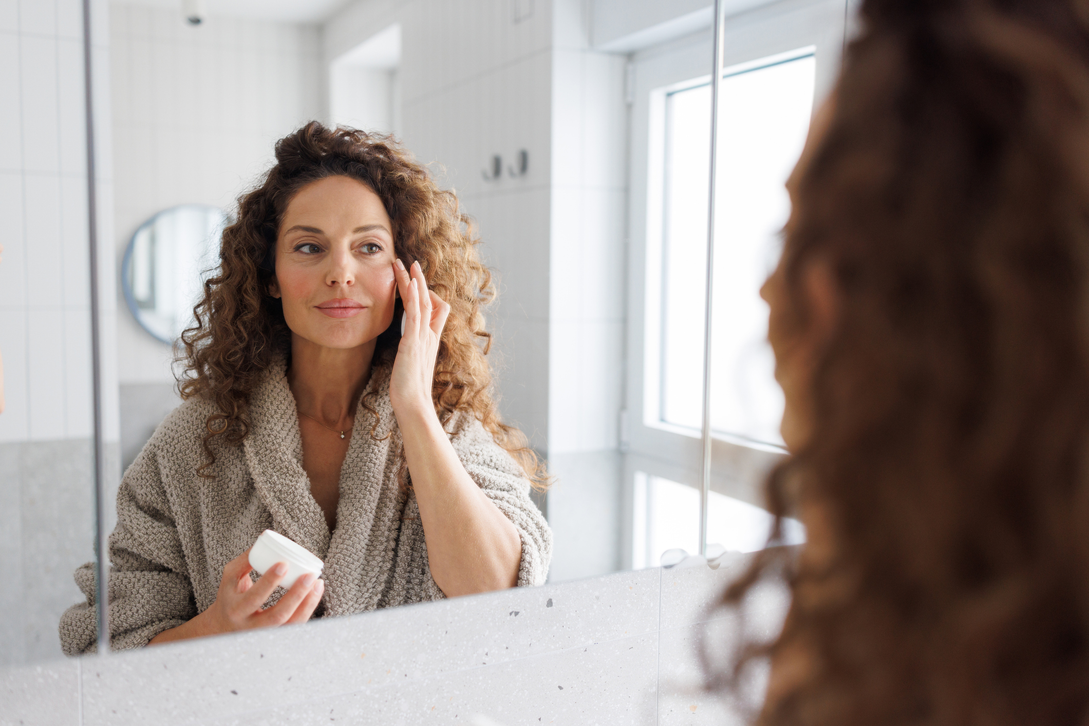 A mid adult woman with curly hair wearing a cozy bathrobe applies face cream to her face in a bright, modern bathroom with natural daylight streaming through the window.