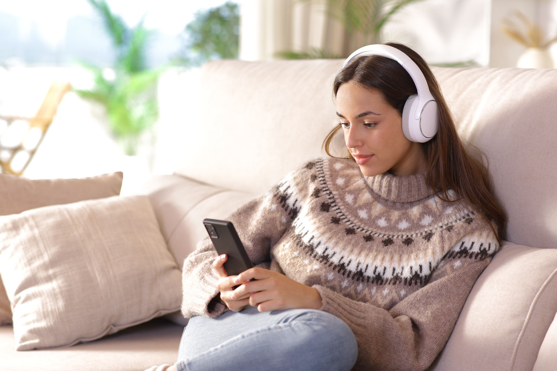 Woman in winter listening audio using phone sitting on a couch at home