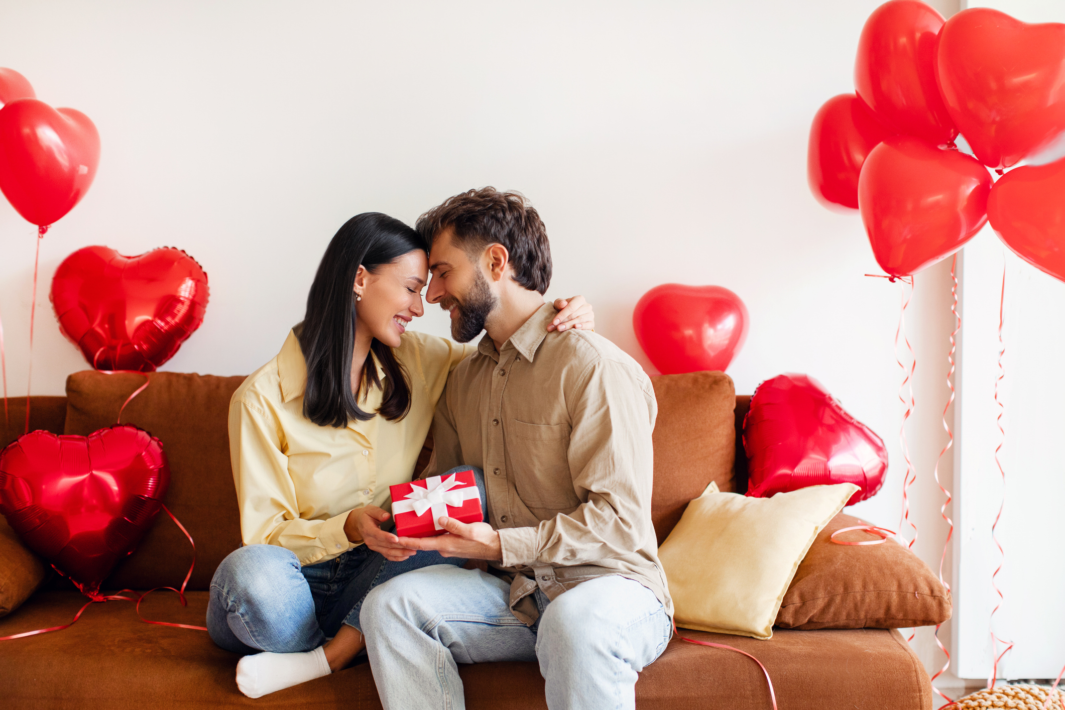 Loving couple embracing, sitting on sofa at home with red heart shape balloons flying around, man and woman bonding, exchanging gifts for Valentine's day