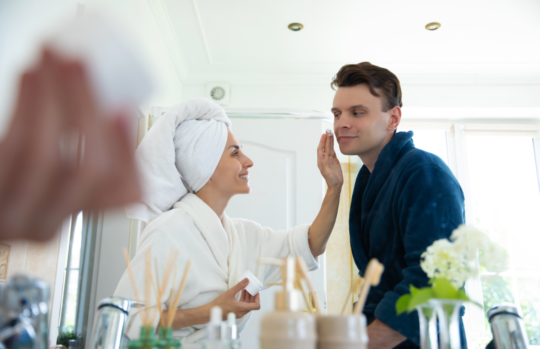 A smiling woman in a bathrobe applies cream to her partners face in a bright bathroom. The couple enjoys a morning skincare routine, sharing a tender, intimate moment in a fresh, modern setting.