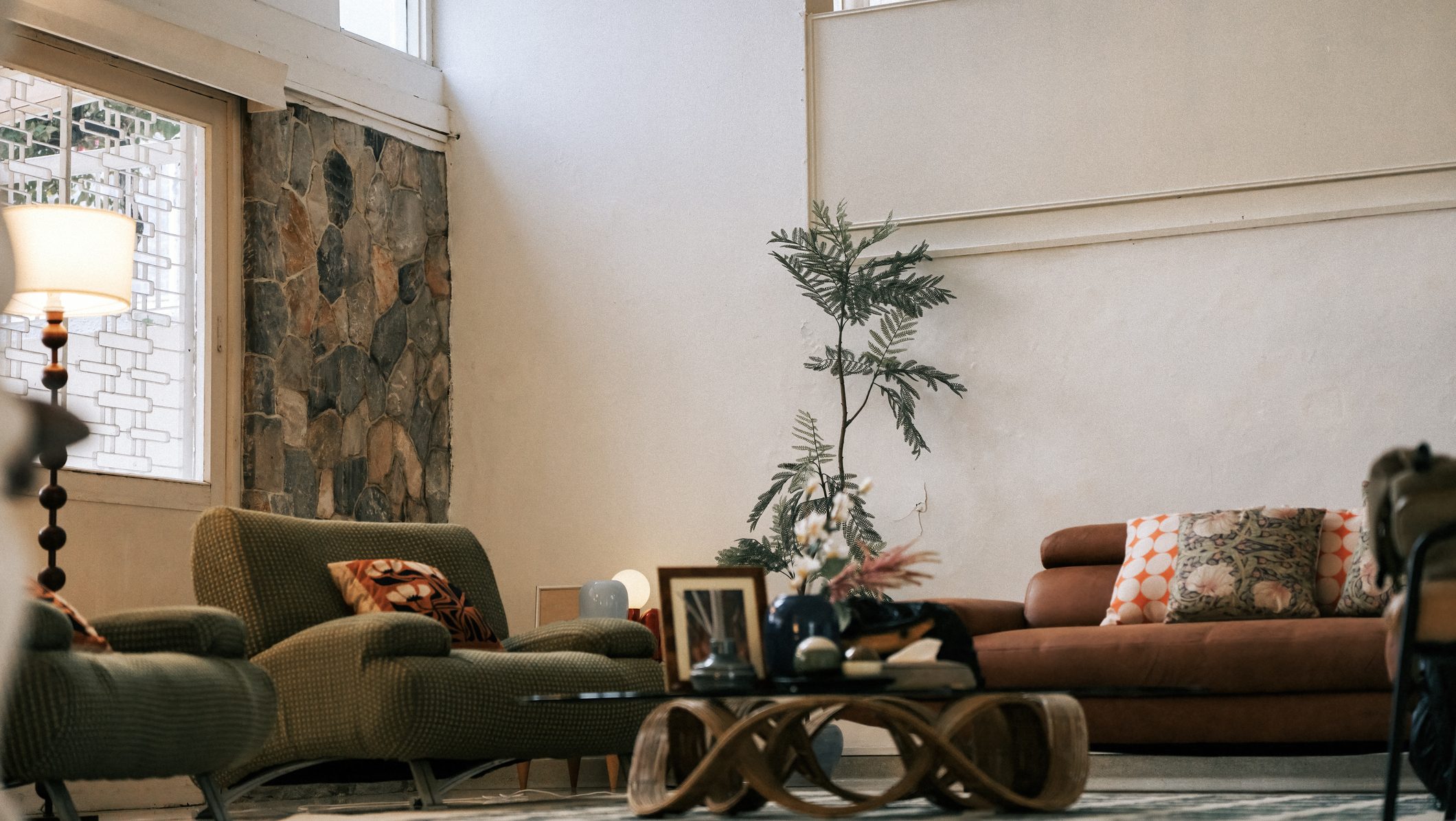 cozy living room featuring mid-century style armchairs, a leather sofa with patterned pillows, a floor lamp, and a stone accent wall, creating a warm and inviting bungalow interior.