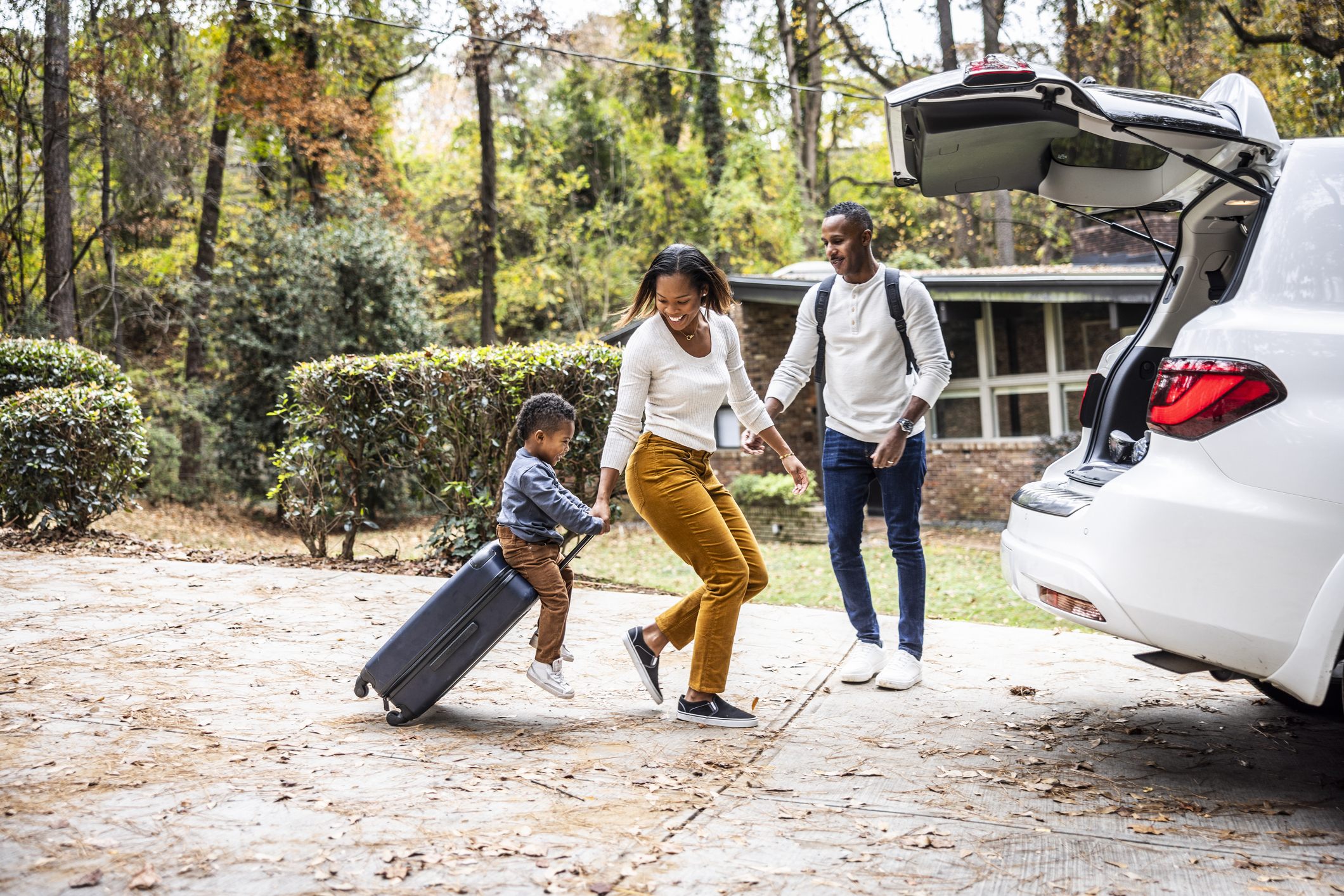 Mother pulling young boy on suitcase and loading car for family vacation