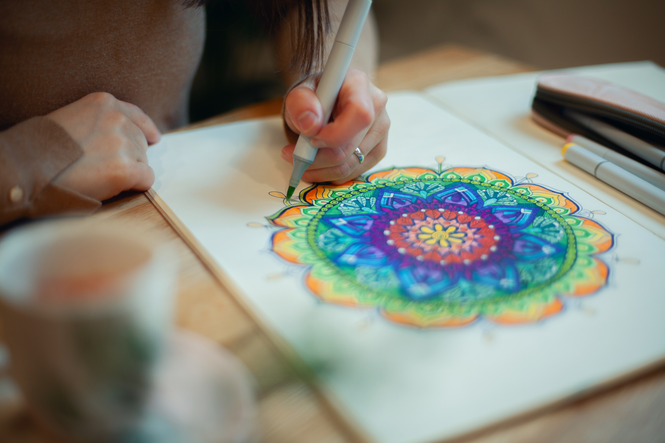 Young woman colouring mandala with Ohuhu alcohol markers and white rosary on table with cup of coffee near flower vase at home. Art therapy and meditation concept. Hobby and leisure during quarantine. Wellbeing.