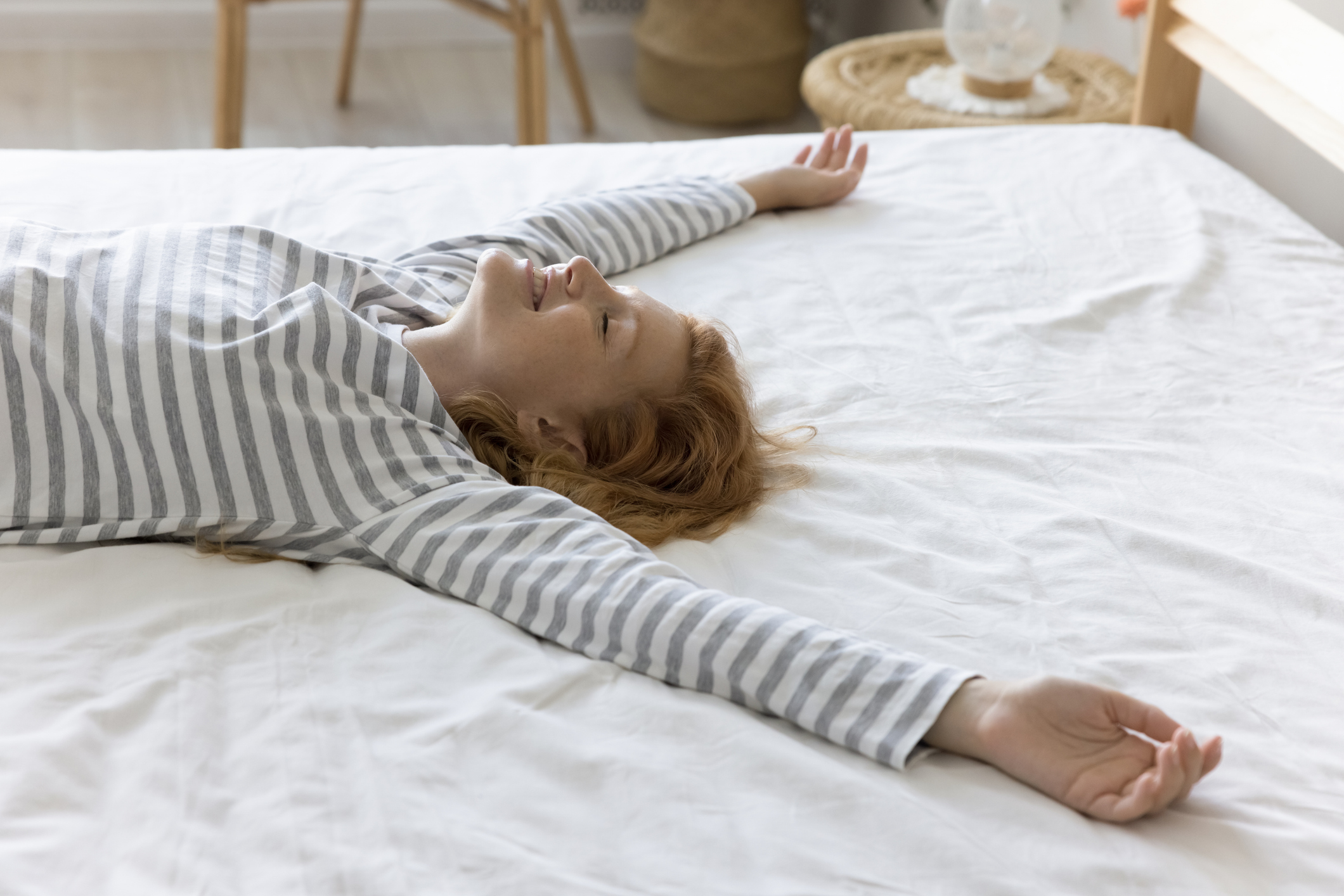 Positive relaxed pretty young woman resting on bed with open arms and closed eyes, lying on back on comfortable soft mattress with white linen, breathing fresh air, relaxing at home