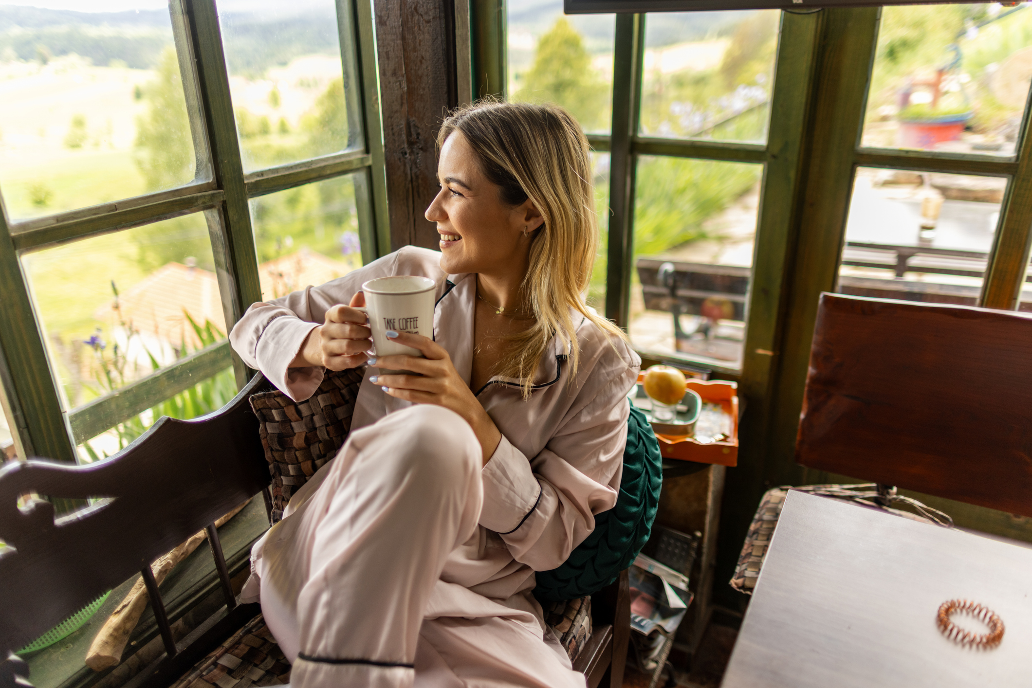 Beautiful mid-adult woman drinking coffee or tea in the morning, wearing pajamas, sitting by the window in a mountain hut looking outside.