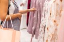 A middle-aged Caucasian woman with short brown hair browses through colorful clothing in a retail store. She is examining pink floral shirts on hangers.