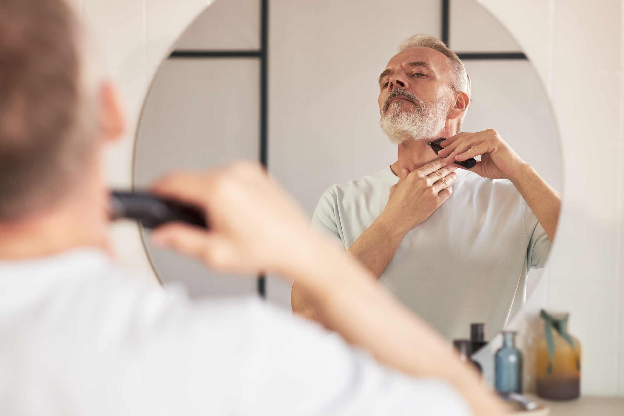 Senior Caucasian man grooming beard with electric trimmer in bathroom, standing in front of mirror and carefully shaping facial hair as part of daily beauty and skincare routine