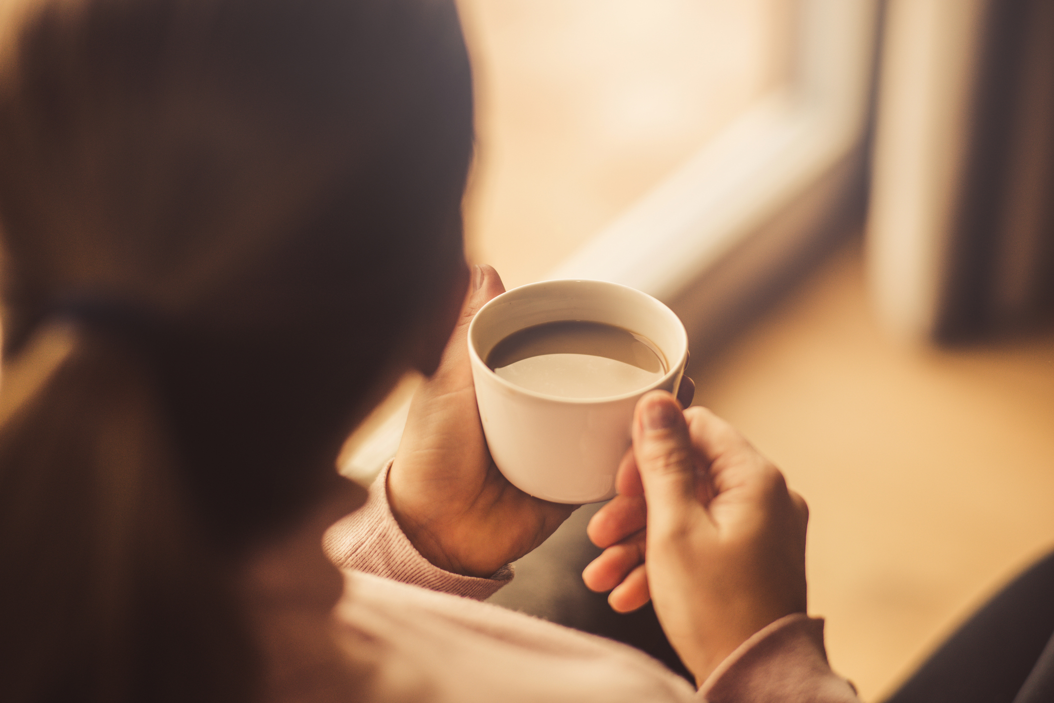 A cup of coffee hold by a woman looking through window.