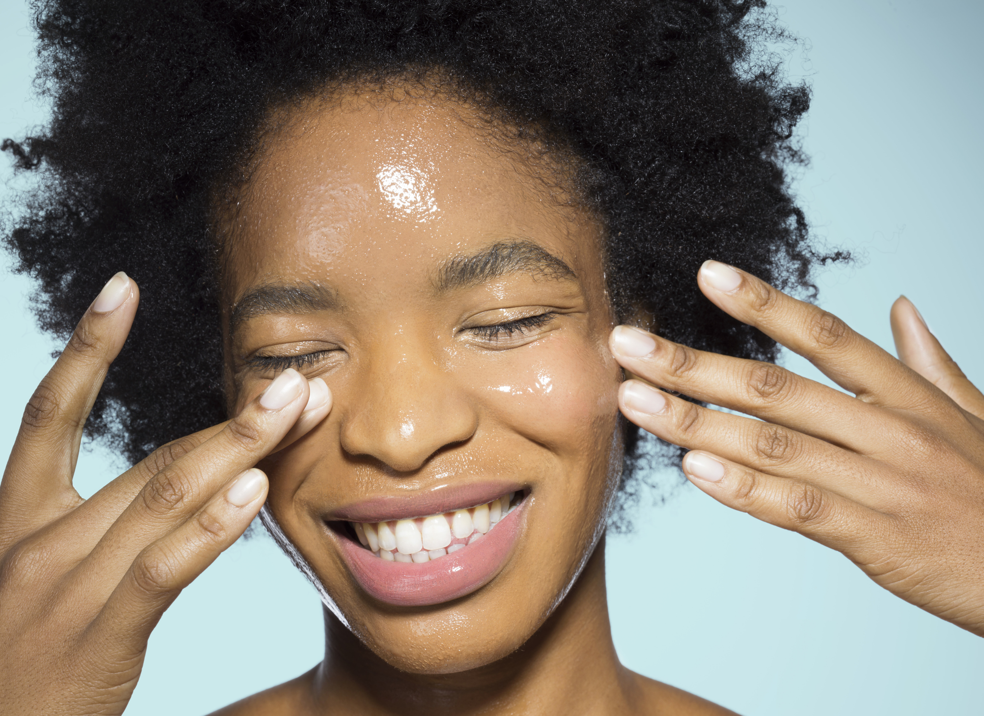 Close-up of young woman applying glossy face make-up moisturizer, smiling, with eyes closed with turquoise background