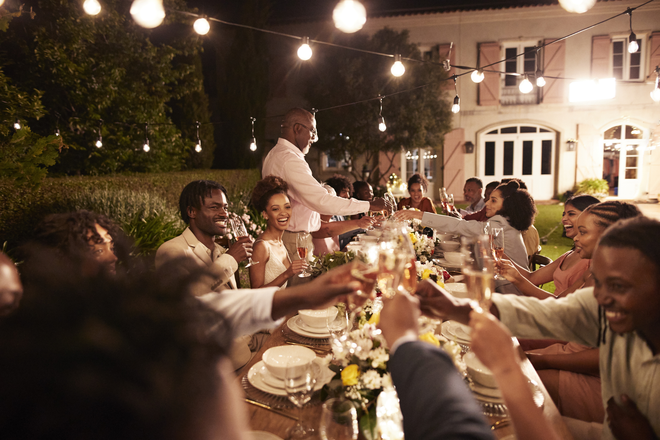 Senior man toasting drinks with guests and newlywed couple during wedding reception dinner party at night under the best outdoor string lights on Amazon