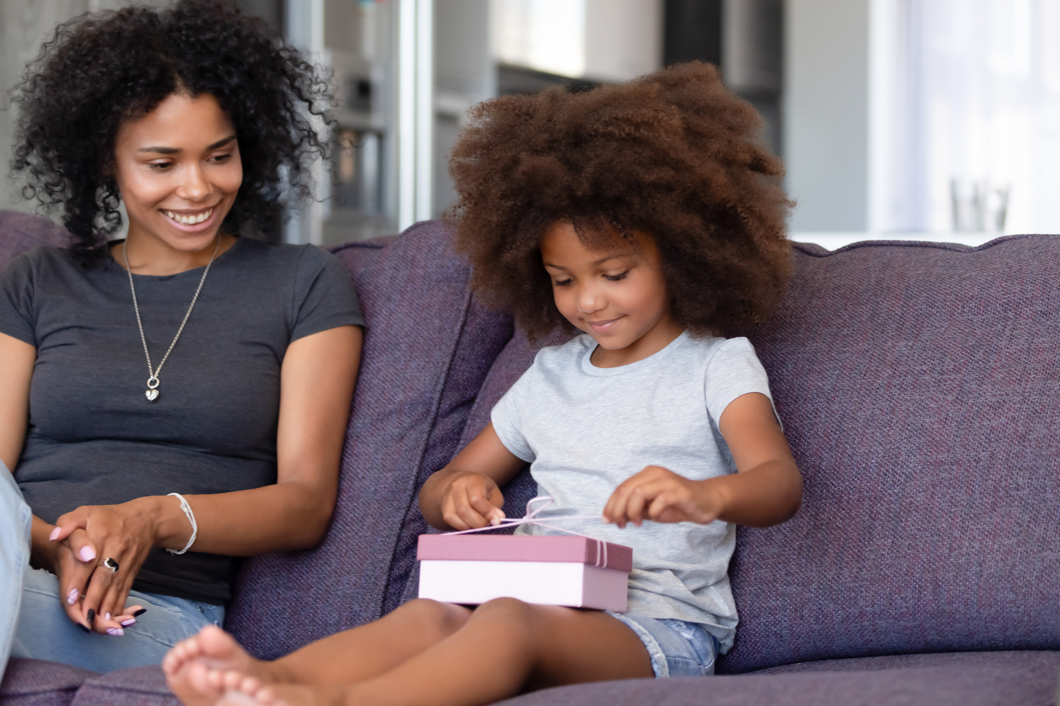 Happy young mom watch small cute daughter unpack birthday gift sitting on couch together, little girl unwrap package presented by mother on special occasion at home