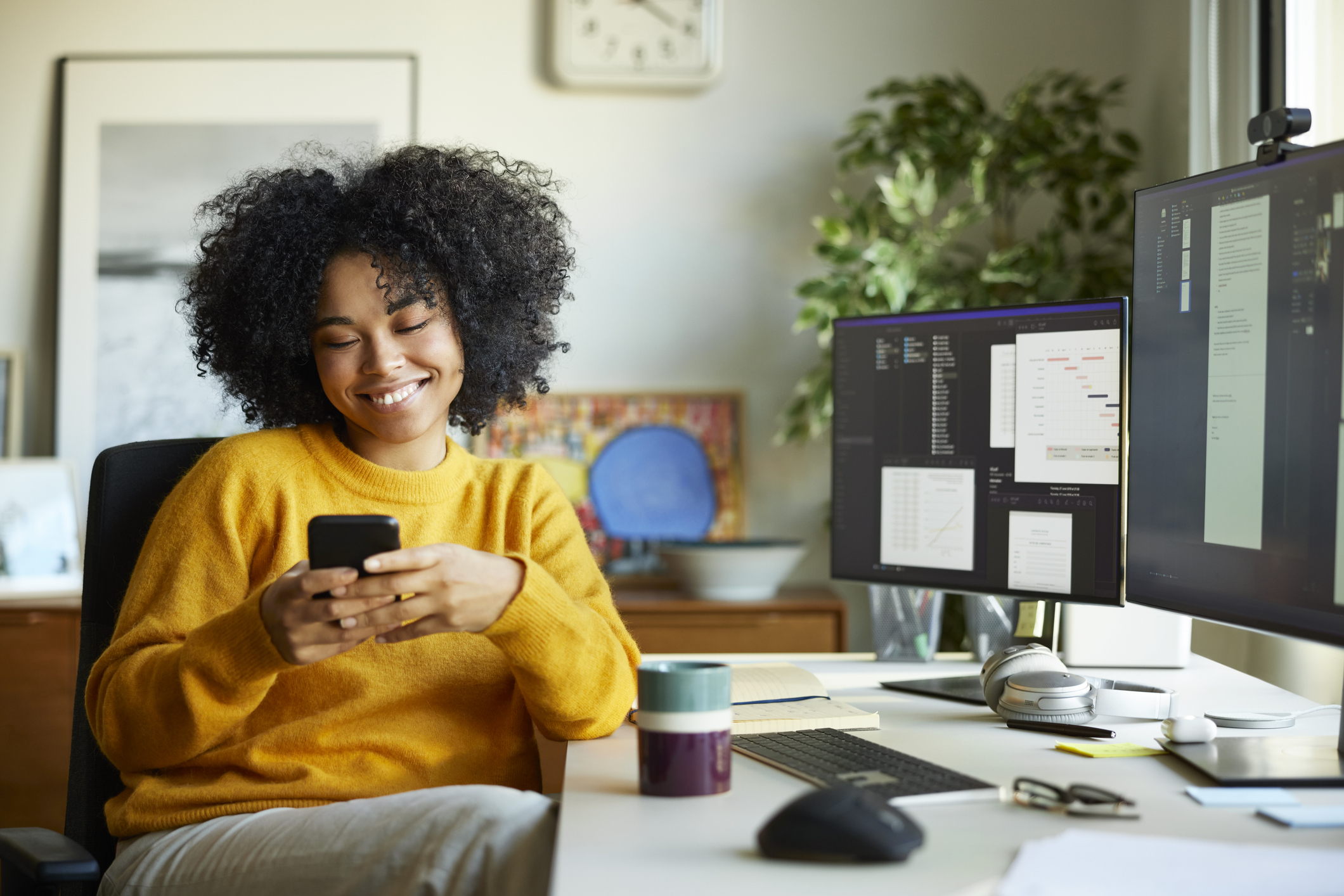 Smiling young businesswoman using smart phone. Young female professional is sitting at computer desk. She is at home office.
