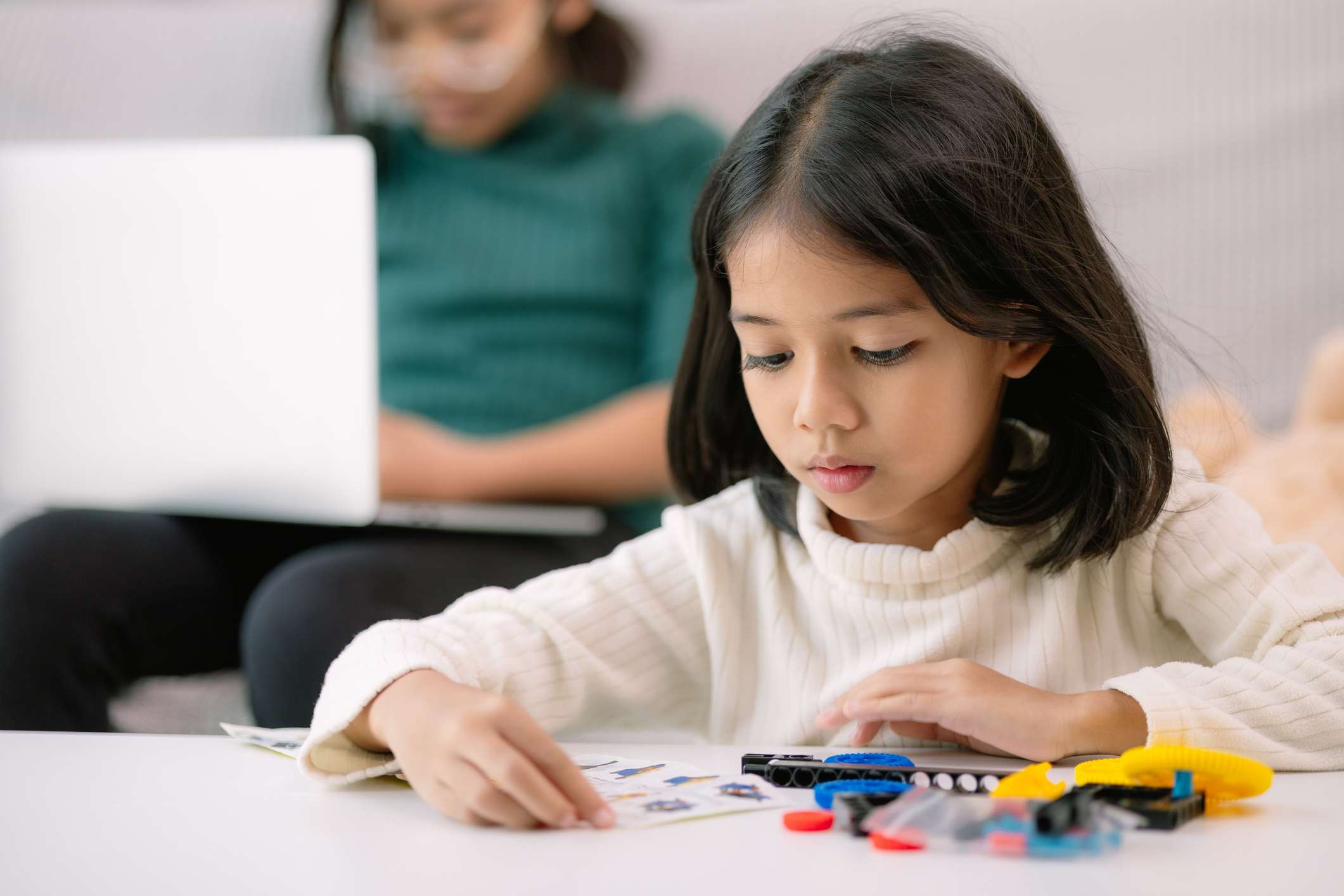 A young girl is sitting at a table with a laptop and a stack of Legos. She is focused on her work, possibly building something with the Legos while using the laptop for reference or inspiration