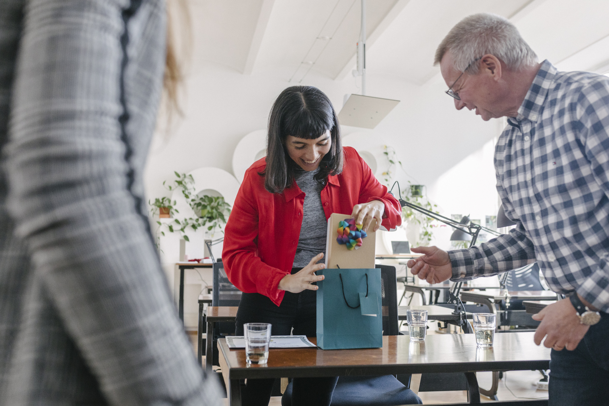 A woman receiving a gift in a bag from her colleagues and celebrating at the office after a recent success.