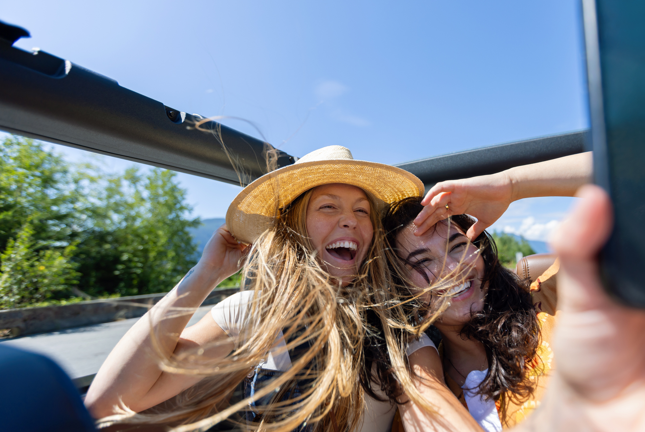 Two female friends sitting in back of convertible car on road trip taking a selfie with mobile phone with long hair blowing in wind, both smiling and enjoying the ride, British Columbia, memories, carefree, fun