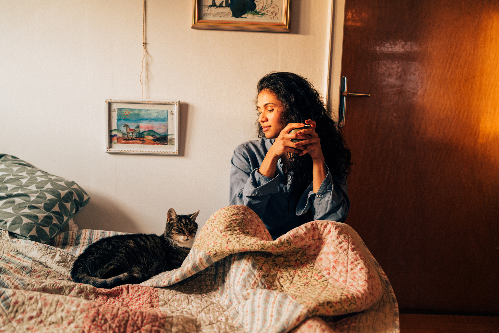 A woman sits on a bed covered with a patterned quilt, gazing thoughtfully while holding a cup. A cat rests nearby, creating a calm indoor moment filled with warmth. Part of a series with video