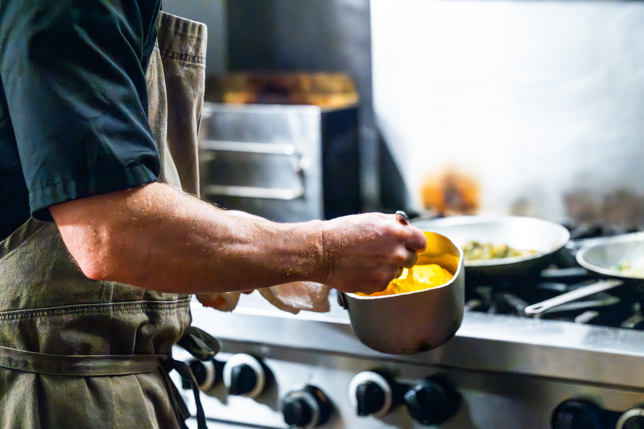 A chef holds a metal bowl with sauce in it in front of a metal gas stovetop. Only the chef's arm and side of his body are in frame as he wears a green apron and a dark shirt. The sauce glows golden in the wooden bowl as the chef dips a spoon into it. In the background greens cook in a pan on a gas stove top.